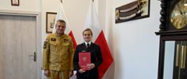 Pomorski komendant wojewódzki Państwowej Straży Pożarnej stoi z funkcjonariuszem kobietą która trzyma czerwoną teczkę za nimi są flagi Polski oraz obraz obok wisi szabla generalska.