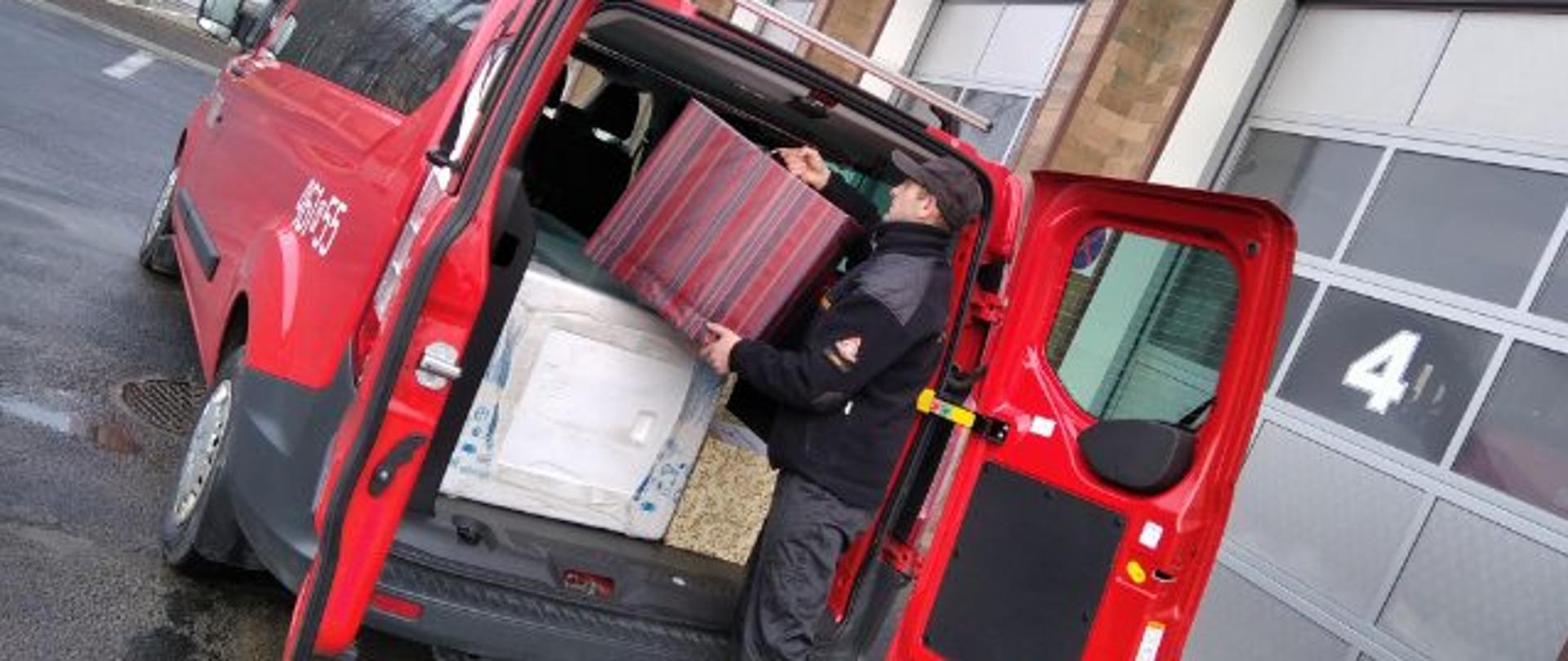 Zdjęcie przedstawia strażaka Jednostki Ratowniczo Gaśniczej Komendy Powiatowej Państwowej Straży Pożarnej w Lesku w trakcie załadunku do busa paczek świątecznych w ramach akcji szlachetna paczka. Na pierwszym planie widać strażaka wkładającego paczkę do samochodu dostawczego który stoi na placu głównym przed budynkiem Komendy Powiatowej Państwowej Straży Pożarnej w Lesku.