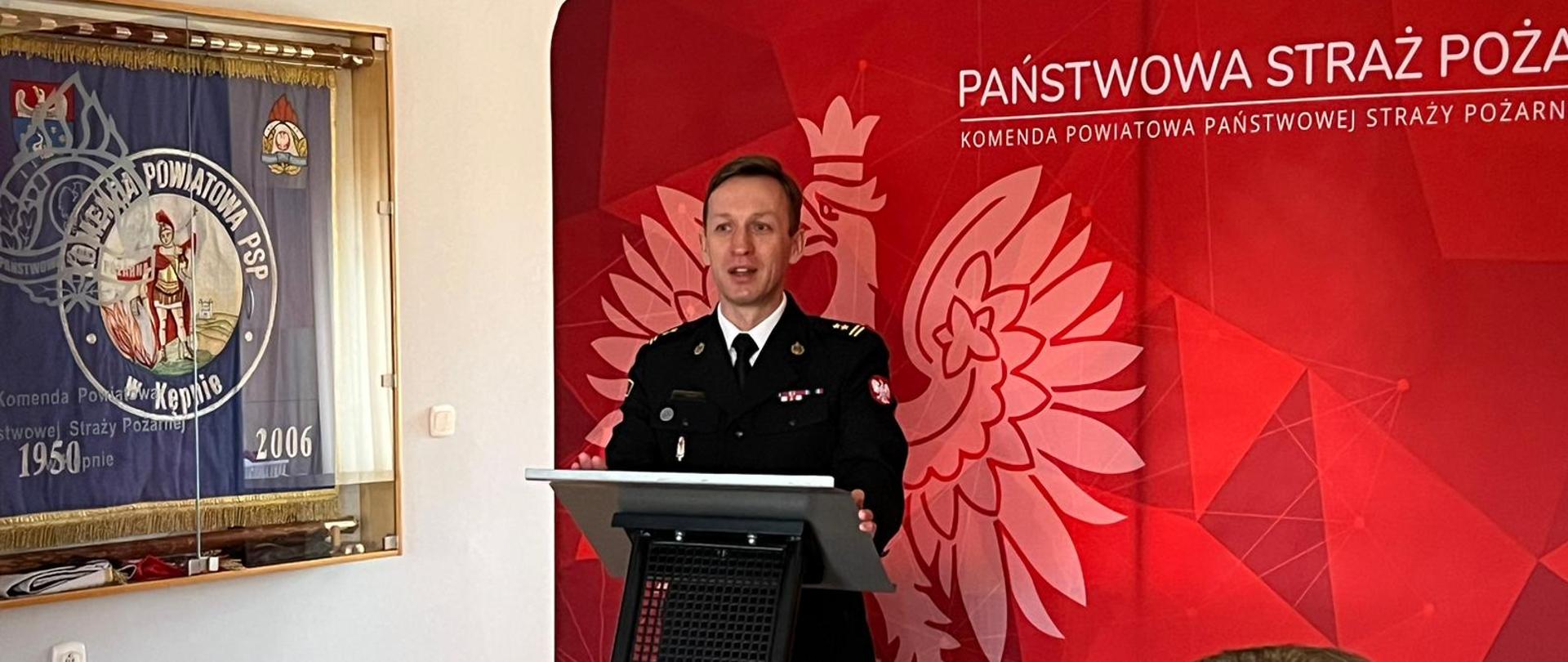 strażak w mundurze stoi przy mównicy, przemawia, za nim stoi czerwona ścianka tekstylna z orłem i napisem państwowa straż pożarna, z lewej strony powieszony sztandar