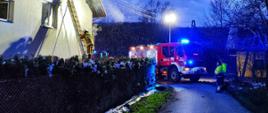 Zdjęcie przedstawia murowany dom w którym pali się dach i poddasze. Widać nad dachem unoszące się kłęby dymu. Do ściany budynku widać przystawiona drabinę. Na drabinie stoi zabezpieczony strażak który ma linie gaśniczą i gasi podbitkę dachu. Po prawej stronie widać czerwony pojazd pożarniczy z włączonymi światłami błyskowymi. Przed pojazdem stoją dwie osoby przedstawiciele pogotowia energetycznego. Samochód pożarniczy ma wysunięty maszt oświetleniowy i oświetla teren działań.