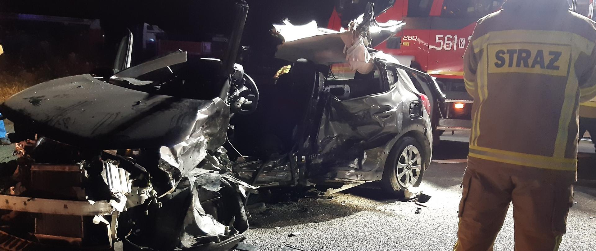 Na zdjęciu samochód osobowy całkowicie zniszczony. Z prawej strony strażak. W dalszej części widoczny samochód ratowniczo - gaśniczy.