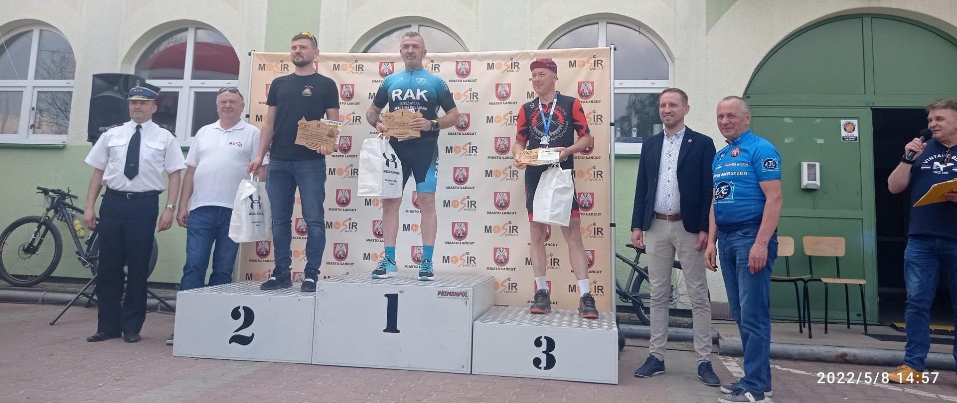 Zdjęcie przedstawia parking przed Miejskim Ośrodkiem Sportu i Rekreacji w Łańcucie. Na podium stoją zawodnicy, którzy zajęli miejsca od 1 do 3 w wyścigu o puchar komendanta powiatowego Państwowej Straży Pożarnej w Łańcucie. Wokół podium stoją: Michał Mglej, zastępca burmistrza miasta Łańcut; Tadeusz Dubiel, prezes Łańcuckiego Towarzystwa Cyklistów; Jerzy Hałka, dyrektor Miejskiego Ośrodka Sportu i Rekreacji oraz st. bryg. Jacek Rejman, zastępca komendanta powiatowego PSP w Łańcucie.
