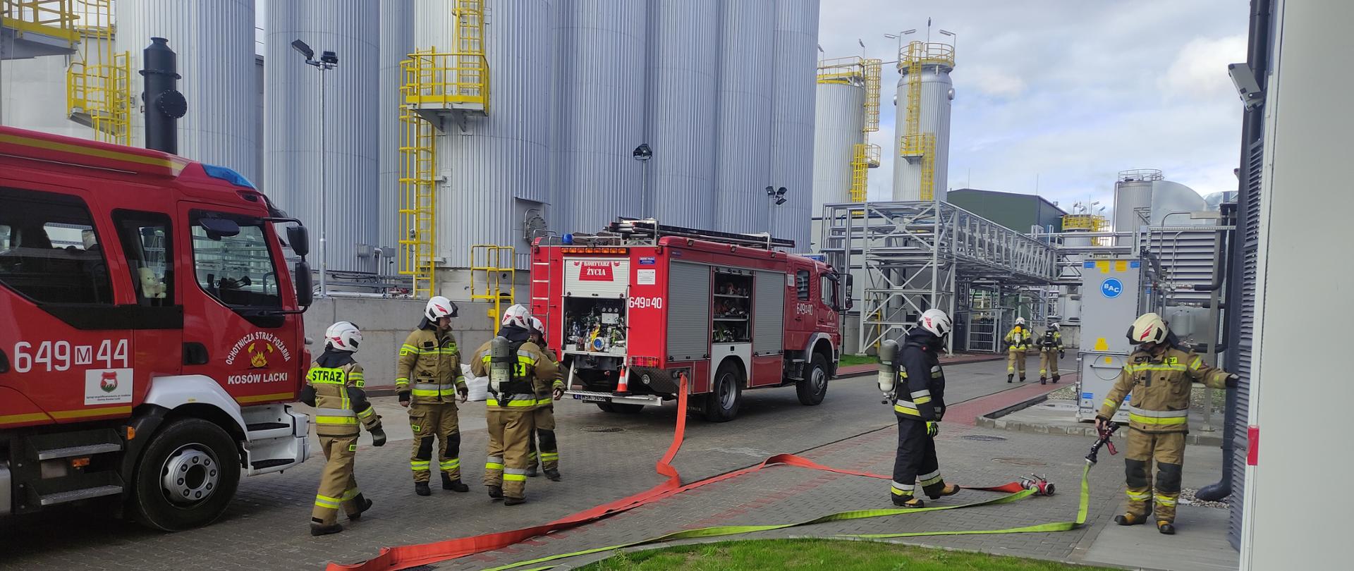 Strażacy rozwijąjący węże strażackie w tle strażacy a za nimi wozy straży pożarnej