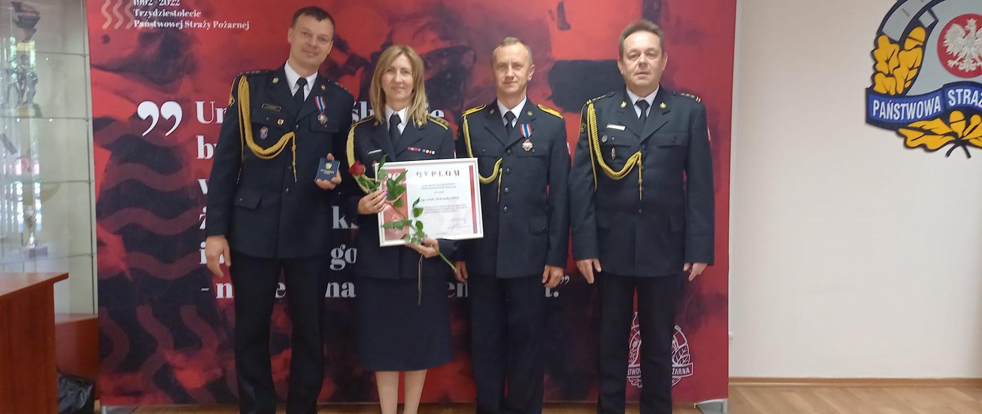 Zdjęcie przedstawia funkcjonariuszy PSP ustawionych przed czerwoną ścianką, fukcjonariusze trzymają w ręku odznaczenia i dyplomy.
