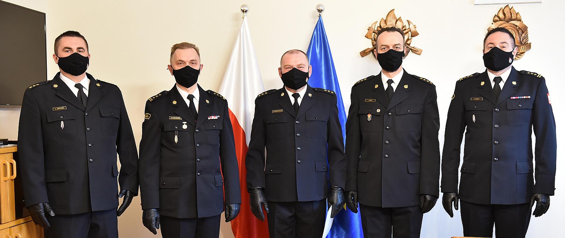 Na zdjęciu pięciu strażaków w umundurowaniu galowym stoją w rzędzie w pomieszczeniu, a nimi na ścianie flaga polski i unii europejskiej oraz dwa drzeworyty, po lewej stronie widać szafkę oraz telewizor po prawej biurko