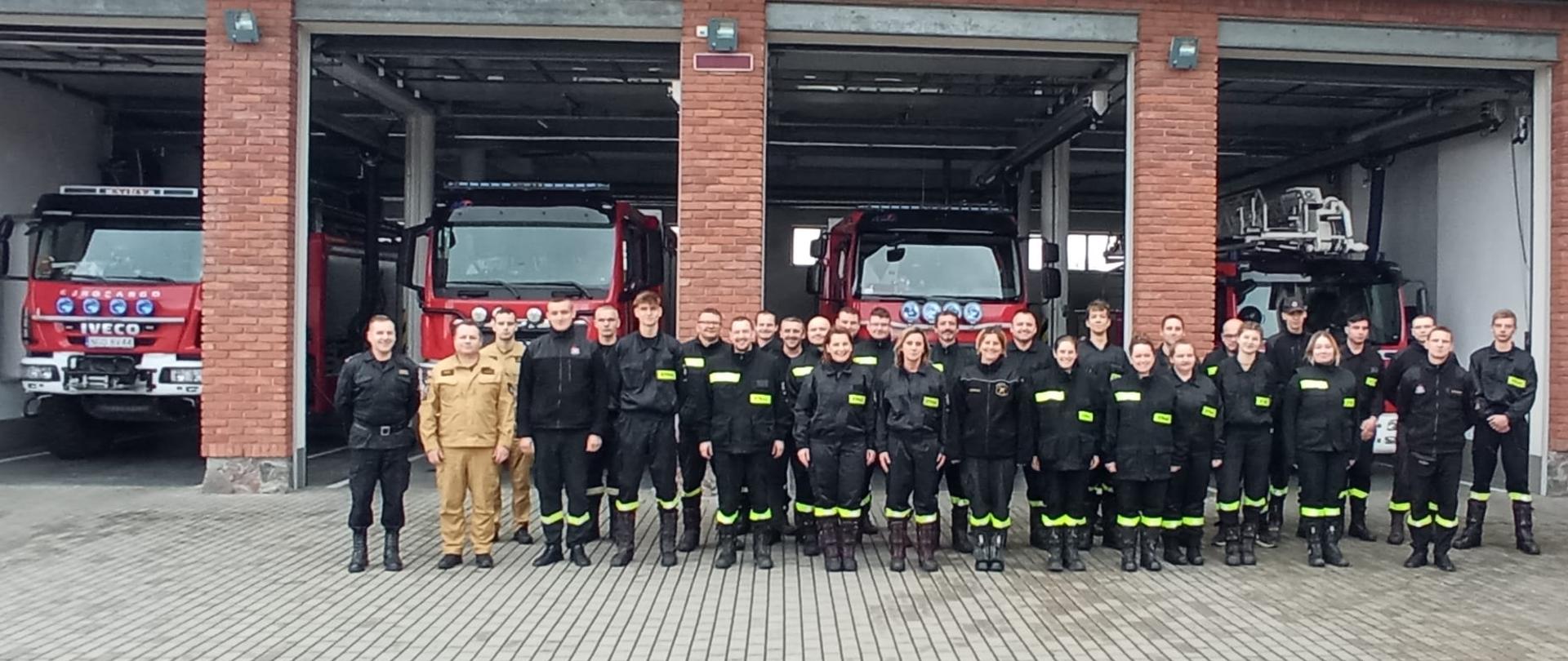 Zdjęcie przedstawia uczestników szkolenia podstawowego strażaka ratownika Ochotniczych Straży Pożarnych, realizowanego w Komedzie Powiatowej Państwowej Straży Pożarnej w Gołdapi wspólnie z komisją egzaminacyjną, na tle pojazdów pożarniczych zlokalizowanych w garażach strażnicy 