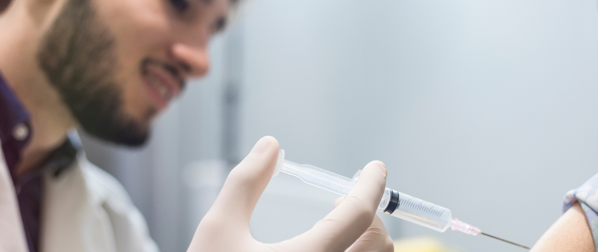 Szczepienie starszej kobiety. Młody lekarz w białym kitlu i rękawiczkach gumowych podaje szczepionkę kobiecie w okularach i szarej bluzce w kwiatki z krótkim rękawem.