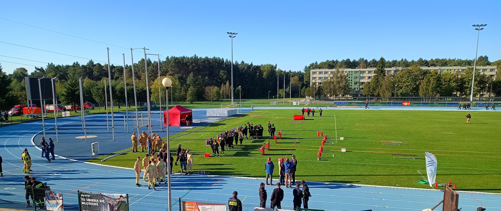 zdjęcie przedstawia stadion oraz drużyny strażaków przygotowujących się do zawodów w oddali bloki i las