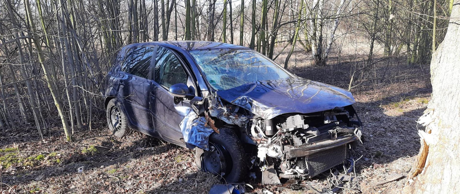 Zdjęcie przedstawia zniszczony samochód osobowy znajdujący się w pasie drogi wojewódzkiej, który brał udział w zdarzeniu. Przed samochodem znajdują się jego uszkodzone części, natomiast za samochodem występuje wysoka roślinność.