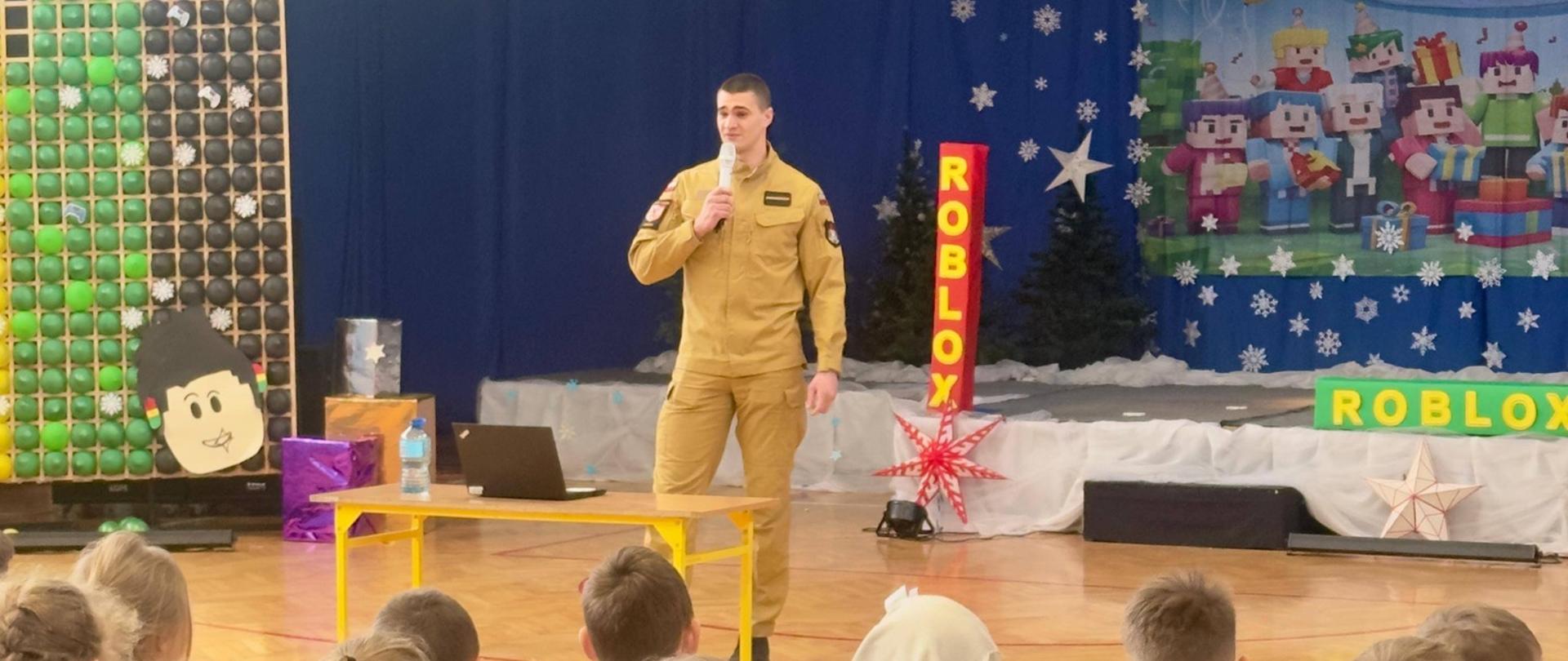 Na zdjęciu widać salę gimnastyczną. Na pierwszym planie siedzi grupa dzieci na krzesełkach, zwróconych twarzami w stronę sceny. Na środku stoi strażak w mundurze, który trzyma mikrofon i prowadzi spotkanie. Obok niego znajduje się niewielki stolik z laptopem. 