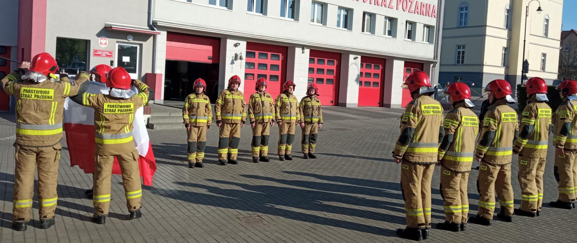 Na zdjęciu strażacy w ubraniach specjalnych w kolorze piaskowym podczas uroczystej zmiany służby, na której podniesiono flagę biało-czerwoną na maszt. Prowadzący zmianę służby w ubraniu dowódczo-sztabowym koloru czarnego. Prowadzący zmianę służby po środku w czarnym umundurowaniu dowódczo-sztabowym.