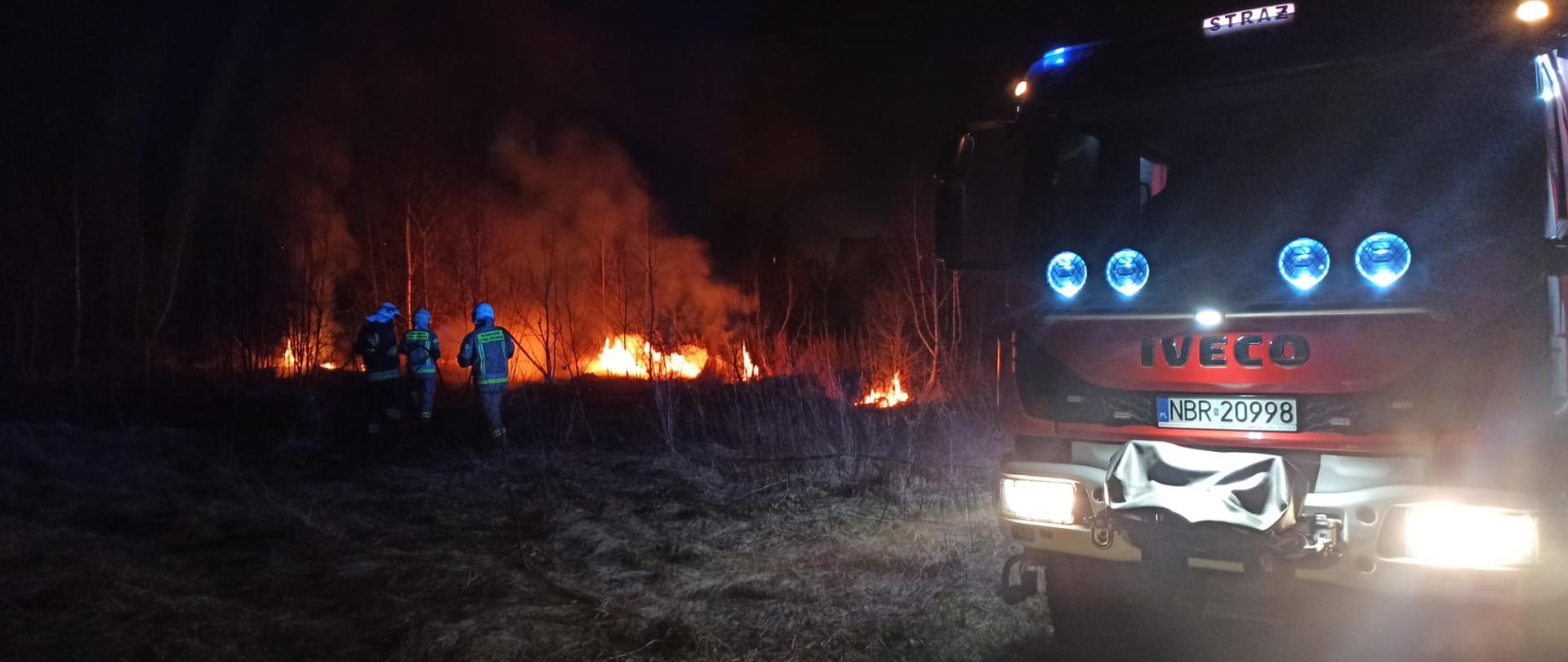 Zdjęcie zrobione nocą. Z prawej strony stoi samochód strażacki na światłach. Dalej trzech strażaków idzie w stronę pożaru. Widać płomienie 