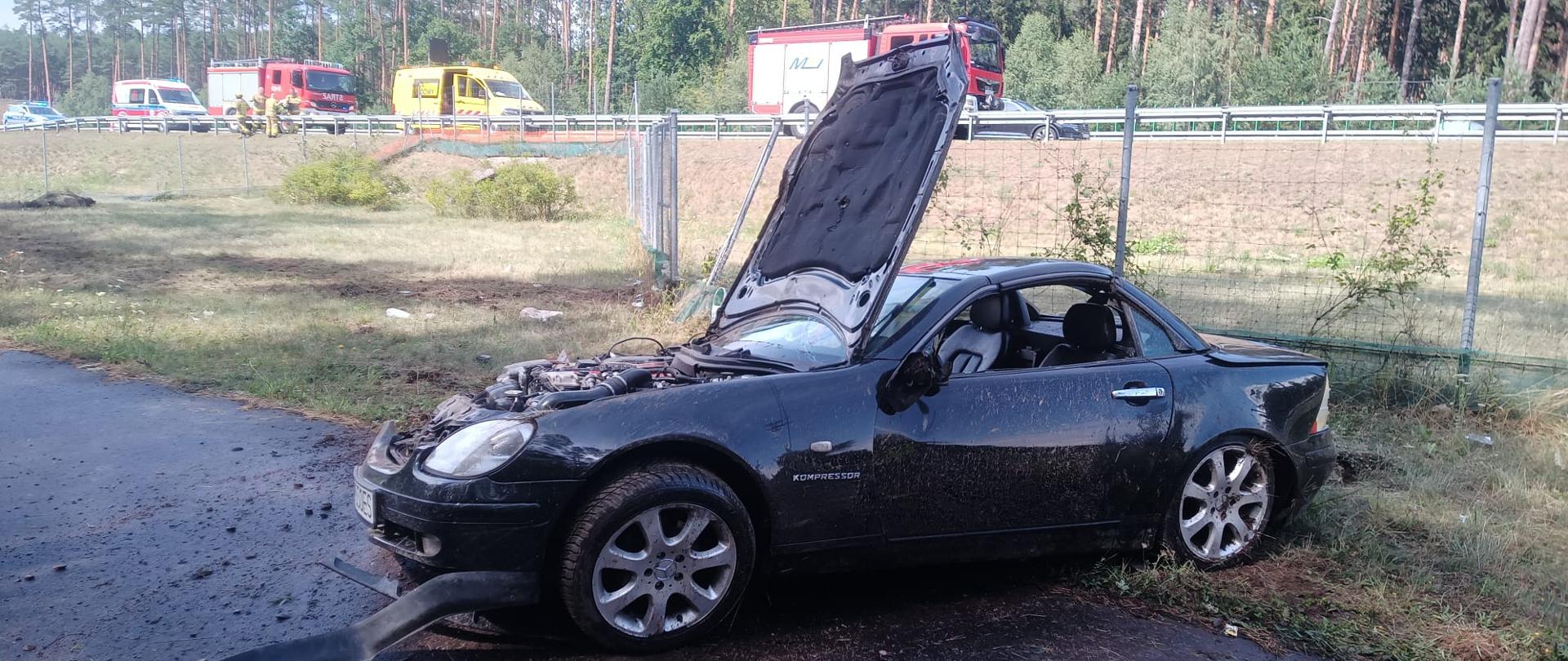 Na zdjęciu widać uszkodzony samochód z podniesioną maską, w tle samochody pożarnicze
