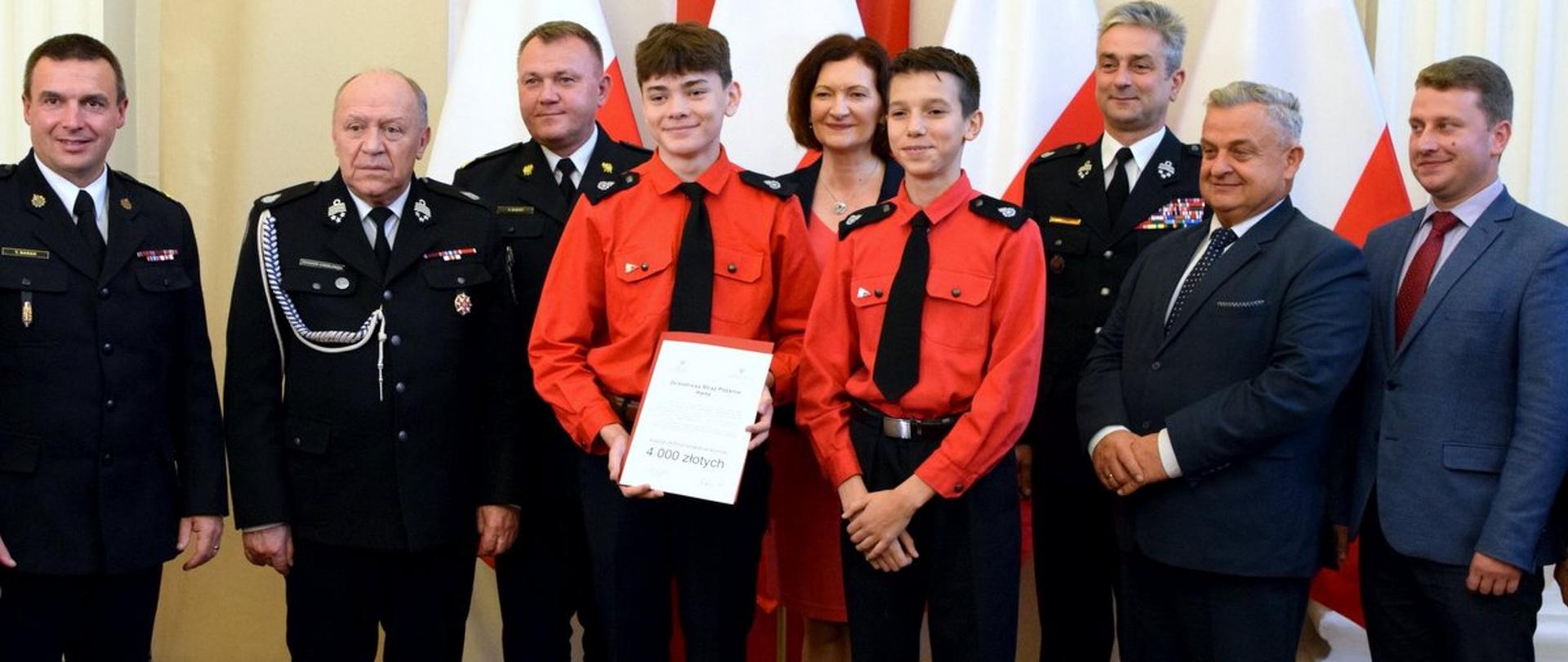 Zdjęcie zrobione w Podkarpackim Urzędzie Wojewódzkim podczas uroczystości. Uczestnicy uroczystości stoją w szeregu, razem z nimi stoją dwaj chłopcy z Młodzieżowej Drużyny Pożarniczej. Młodzi druhowie trzymają promesę. 