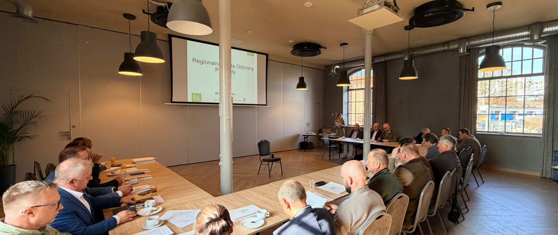 Duży stół, na którym leżą dokumenty oraz stoją filiżanki z kawą/herbatą. Wokół stołu siedzą ludzie i patrzą na wyświetlaną prezentację. 