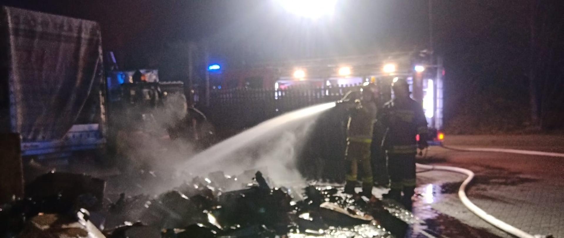 Strażacy przelewają wodą towar wyniesiony z pożaru obiektu magazynowego.