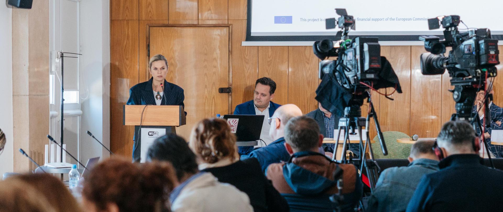 Kobieta przemawia z mównicy do publiczności. Przed nią stoją duże kamery na statywach. W tle widoczny jest ekran z napisem "25 - 26 September 2025".