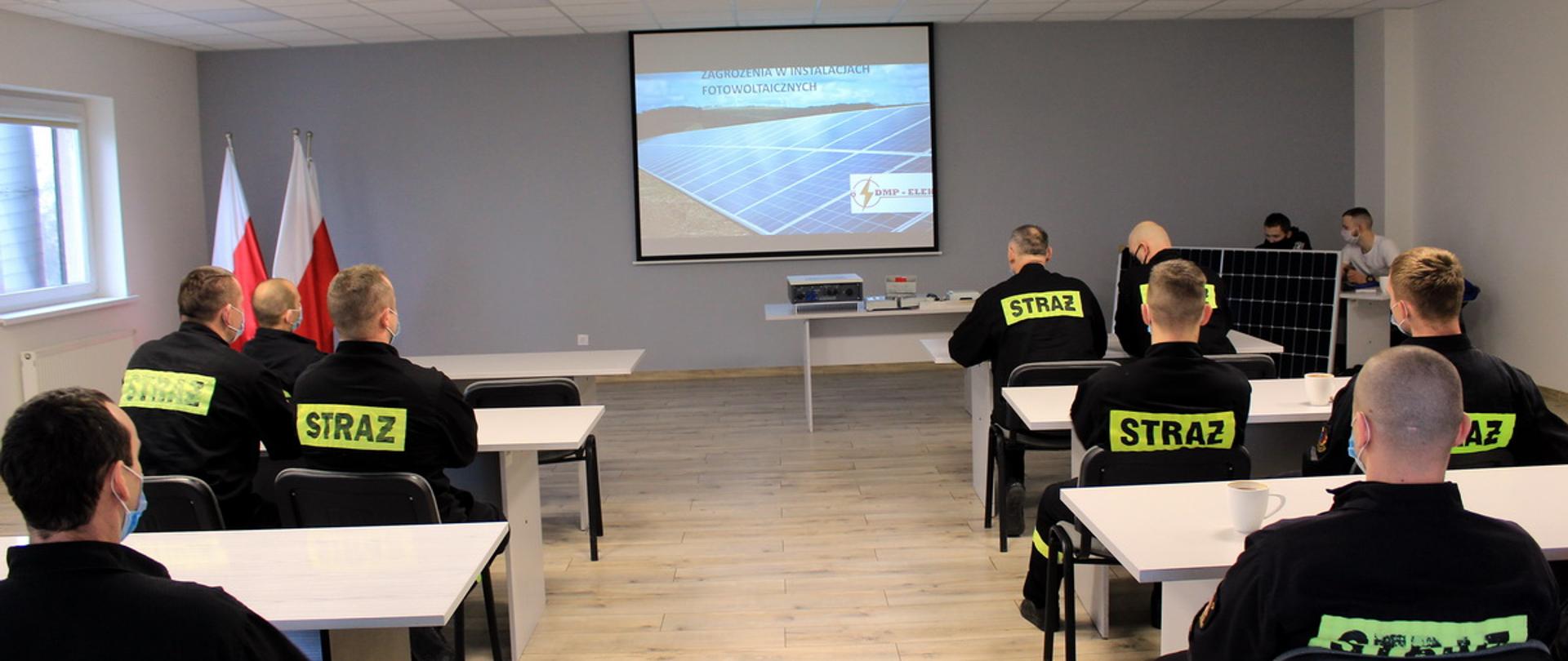 Zdjęcie Funkcjonariuszy Państwowej Straży Pożarnej uczestniczący w szkoleniu dotyczącym instalacji fotowoltaicznej. W centrum zdjęcia widać duży ekran, na którym wyświetlana jest prezentacja multimedialna. Po prawej i lewej stronie widoczni są strażacy biorący udział w szkoleniu. 
