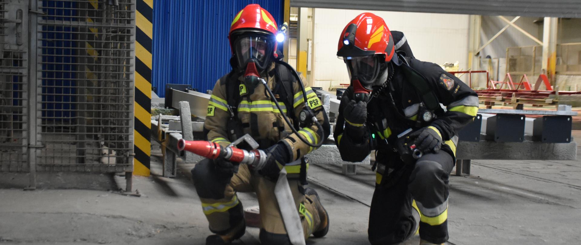 Zdjęcie przedstawia wnętrze hal produkcyjnych. Dwóch strażaków w pełnym umundurowaniu specjalnym oraz pracujący w aparatach ochrony dróg oddechowych udający się do strefy zagrożenia.