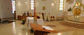 Na zdjęciu widoczny ołtarz w kościele. Widzimy również księdza odprawiającego mszę św. w oddali widoczni pozostali księża i ministranci. Wszyscy księża ubrani w białe ornaty.