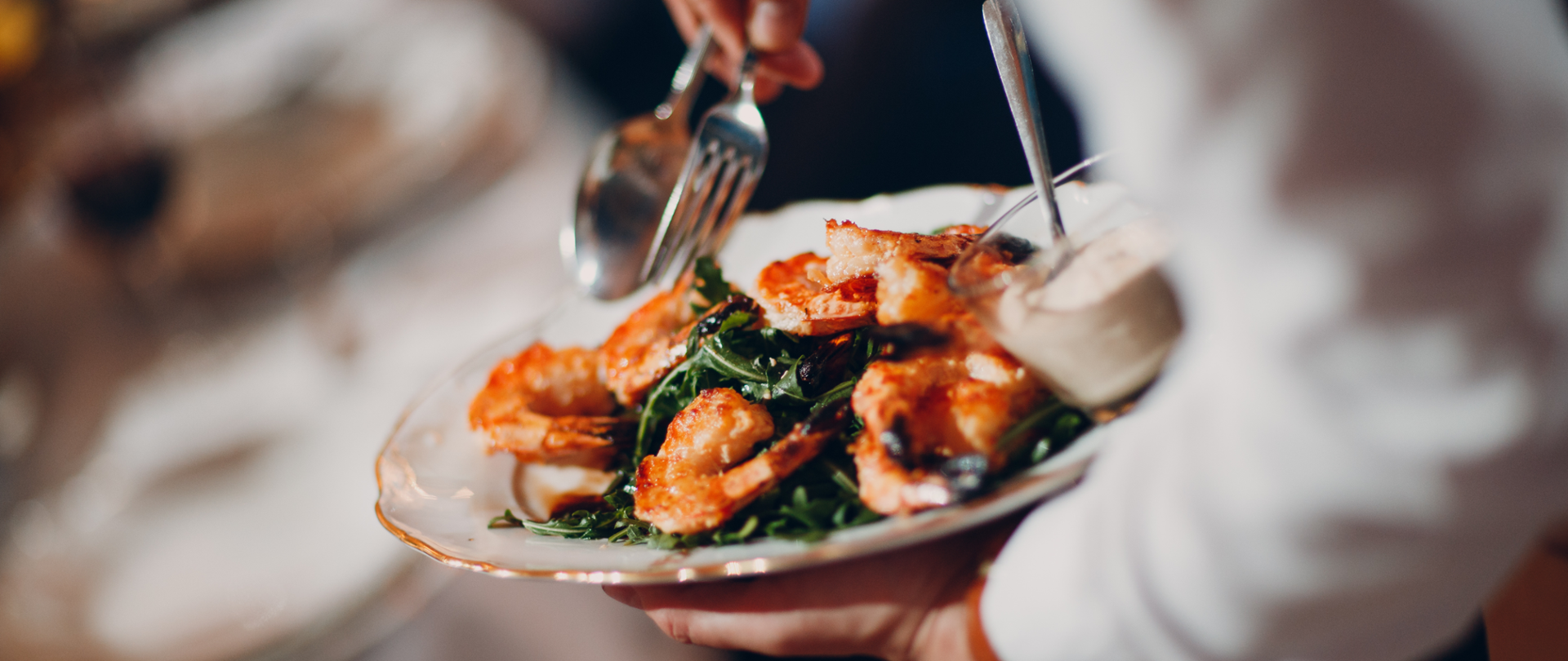 Na zdjęciu widać rękę w białej koszuli trzymającą talerz. Na talerzu znajdują się krewetki i liście sałaty oraz sos w słoiczku.