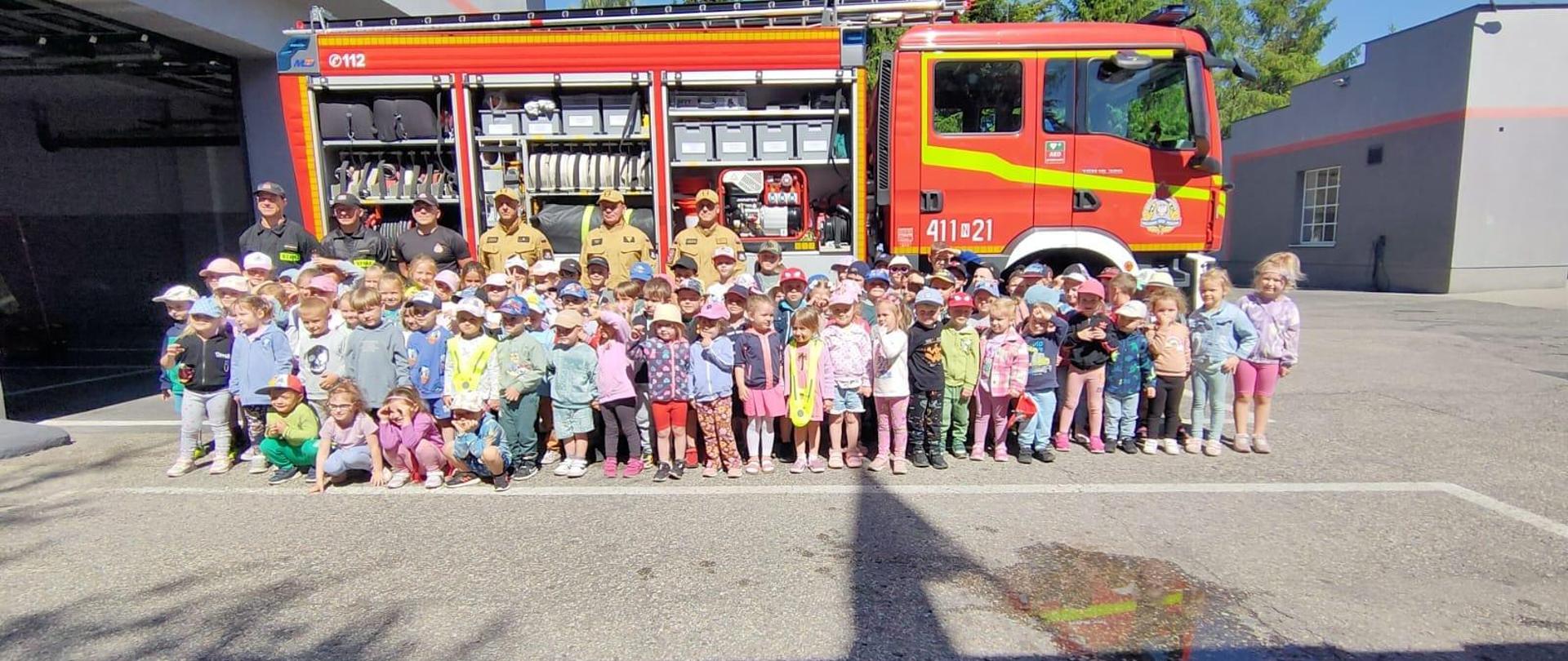 Na zdjęciu duża grupa dzieci z przedszkola. Stoją ze strażakami na tle dużego samochodu strażackiego. Słonecznie.