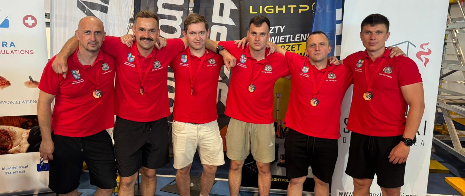 Fotografia przedstawia funkcjonariuszy ubranych w czerwone koszulki i czarne spodenki stojących razem po dekoracji. Na szyjach udekorowani okolicznościowymi medalami. 