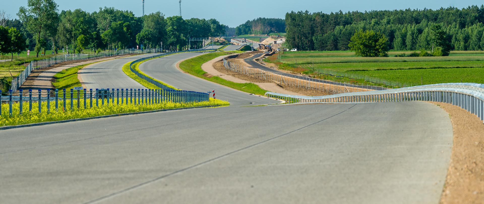 Na zdjęciu widoczna jest budowana droga ekspresowa, w tle widać drzewa i pracujące maszyny budowlane.