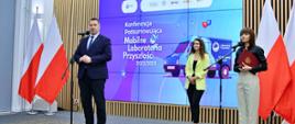 Na sali stoi minister Czarnek i mówi do mikrofonu na stojaku, obok niego dwie kobiety, za nimi wielki ekran z napisem Konferencja podsumowująca Mobilne Laboratoria Przyszłości.