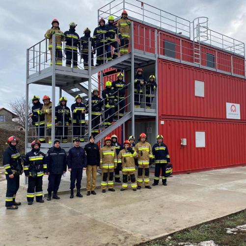 Grupa 26 osób ubranych w stroje strażackie pozuje do zdjęcia przy czerwonym trenażu zbudowanym z kontenerów.