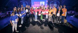 Grupa kolorowo ubranej młodzieży stoi na czarnej podłodze, w ciemnym pomieszczeniu, za nimi duży świecący na niebiesko i fioletowo monitor z napisem HackEdu.