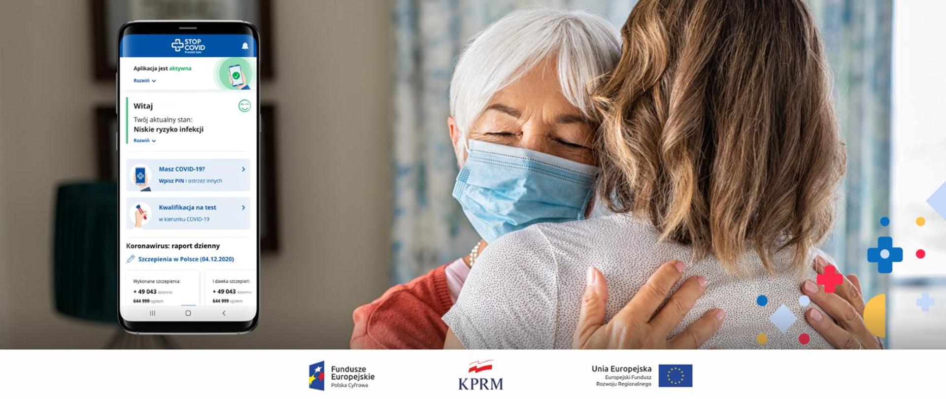 Zdjęcie, na którym widoczne są dwie osoby, które się przytulają. Tuż obok widnieje telefon komórkowy, na którego ekranie widoczna jest aplikacja STOP COVID.