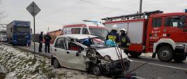 Na zdjęciu widać uszkodzony pojazd marki Daewoo Matiz który znajduje się na poboczu drogi, samochód gaśniczy jednostki OSP oraz karetkę pogotowia. Dwóch ratowników straży pożarnej usuwa skutki zdarzenia. Dwóch funkcjonariuszy Policji prowadzi czynności wyjaśniające zdarzenie.