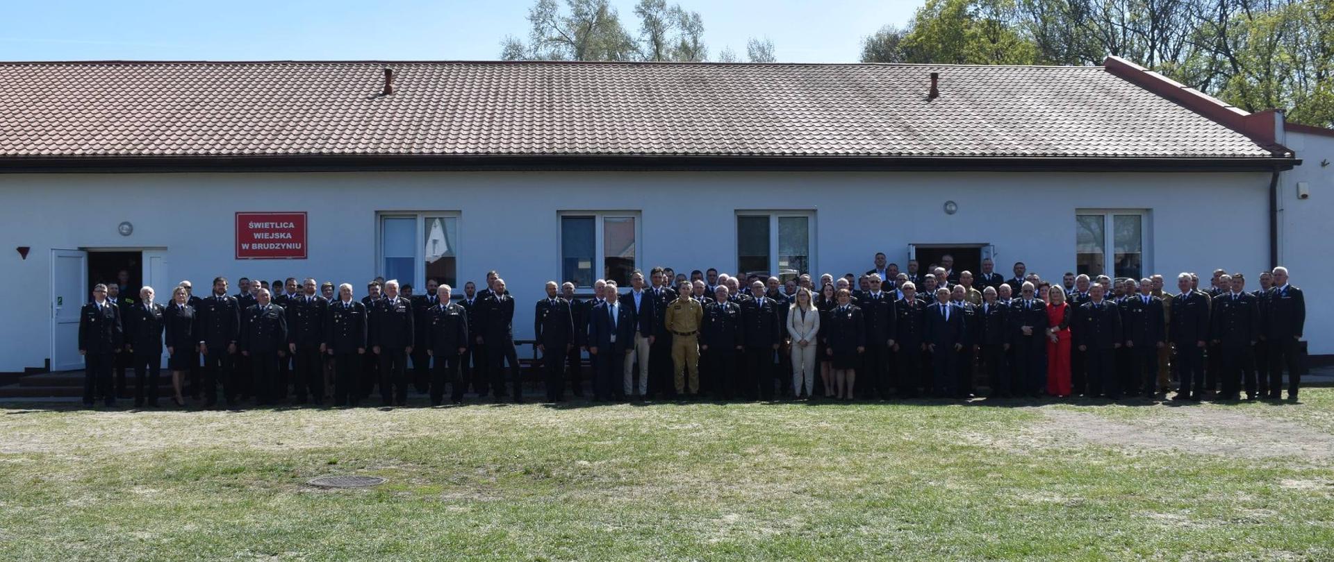 Duża grupa osób, głównie w mundurach, stoi ustawiona w rzędzie przed długim, jasnym budynkiem z czerwonym dachem. Zdjęcie wykonano na zewnątrz, na trawiastym terenie, w słoneczny dzień.
