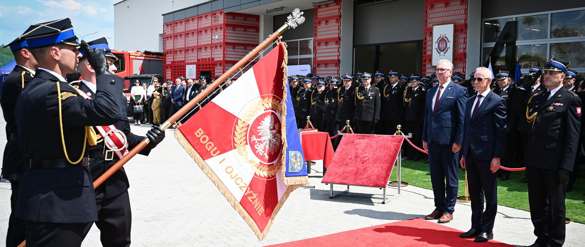 Poczet sztandaru prezentuje nowo nadany sztandar Komendy Powiatowej PSP w Brzozowie. Trzech strażaków w asyście salutuje na czerwonym dywanie. Widoczna jest czerwona strona sztandaru z godłem Polski (Orłem Białym) w wieńcu oraz haftowanym złotym napisem „BOGU I OJCZYŹNIE”. Naprzeciwko stoją oficjele.