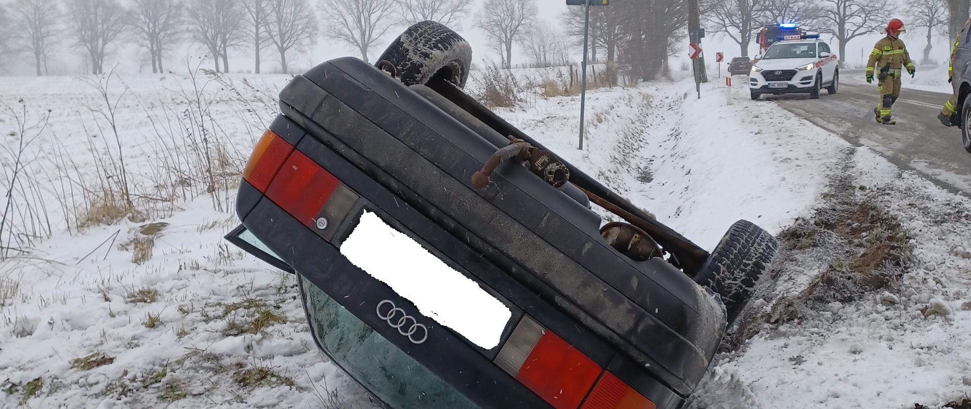 Samochód osobowy znajduje się w rowie na dachu. Obok pojazdu leży fotelik. W oddali widać samochody PSP oraz strażaków. 