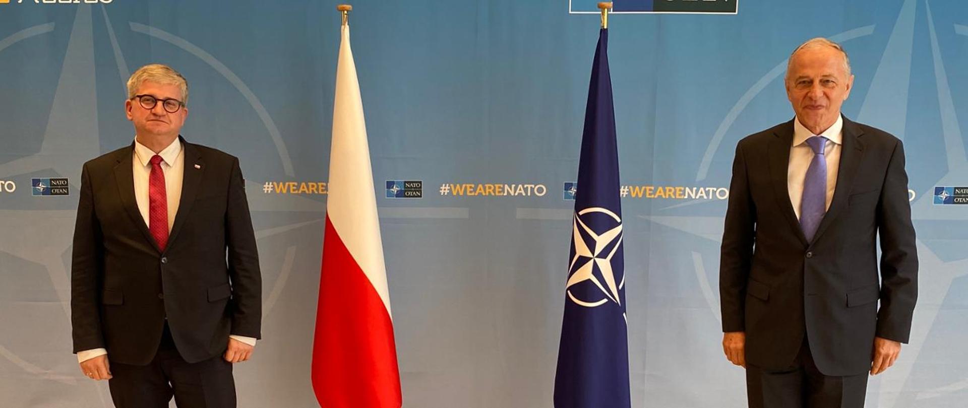 Minister Soloch i Zastępca Sekretarza Generalnego stoją na tle flag Polski i NATO