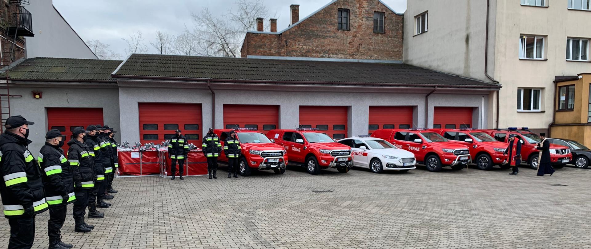 Zdjęcie obrazujące strażaków ustawionych w dwuszeregu w umundurowaniu dowódczo-sztabowym i kapelani strażaccy w szatach liturgicznych dokonujący poświęcenia pojazdów i sprzętu pożarniczego podczas uroczystego przekazania.