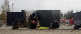 Na zdjęciu strażacy w ubraniach specjalnych oraz aparatach oddechowych po wyjściu z komory ogniowej. W tle komora oraz wydobywający się z niej dym
