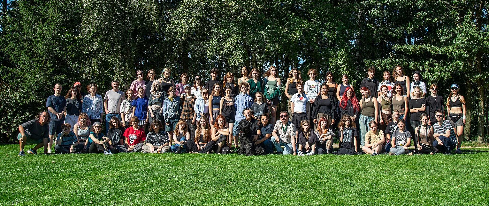 Zdjęcie przedstawia dużą grupę osób pozujących do wspólnej fotografii na świeżym powietrzu. Ludzie stoją i siedzą w rzędach na zielonej trawie. W tle znajduje się las z dużymi drzewami. Wszyscy są swobodnie ubrani i uśmiechają się do aparatu, sprawiając wrażenie zrelaksowanych i szczęśliwych.