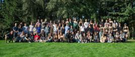 Zdjęcie przedstawia dużą grupę osób pozujących do wspólnej fotografii na świeżym powietrzu. Ludzie stoją i siedzą w rzędach na zielonej trawie. W tle znajduje się las z dużymi drzewami. Wszyscy są swobodnie ubrani i uśmiechają się do aparatu, sprawiając wrażenie zrelaksowanych i szczęśliwych.