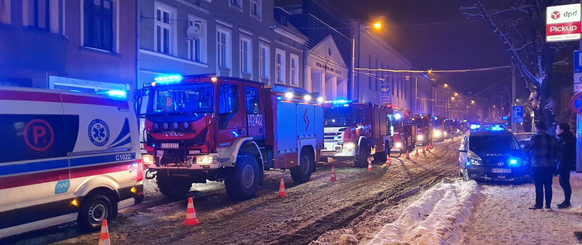 Zdjęcie przedstawia ulicę w centrum miasta nocną porą. W świetle latarni ulicznych widoczne pojazdy Pogotowia Ratunkowego i Straży Pożarnej stojące w rzędzie na jednym pasie ulicy, oraz policyjny radiowóz stojący z drugiej strony. W tle fasady kamienic. Na powierzchni ulicy i poboczu zalega warstwa śniegu.