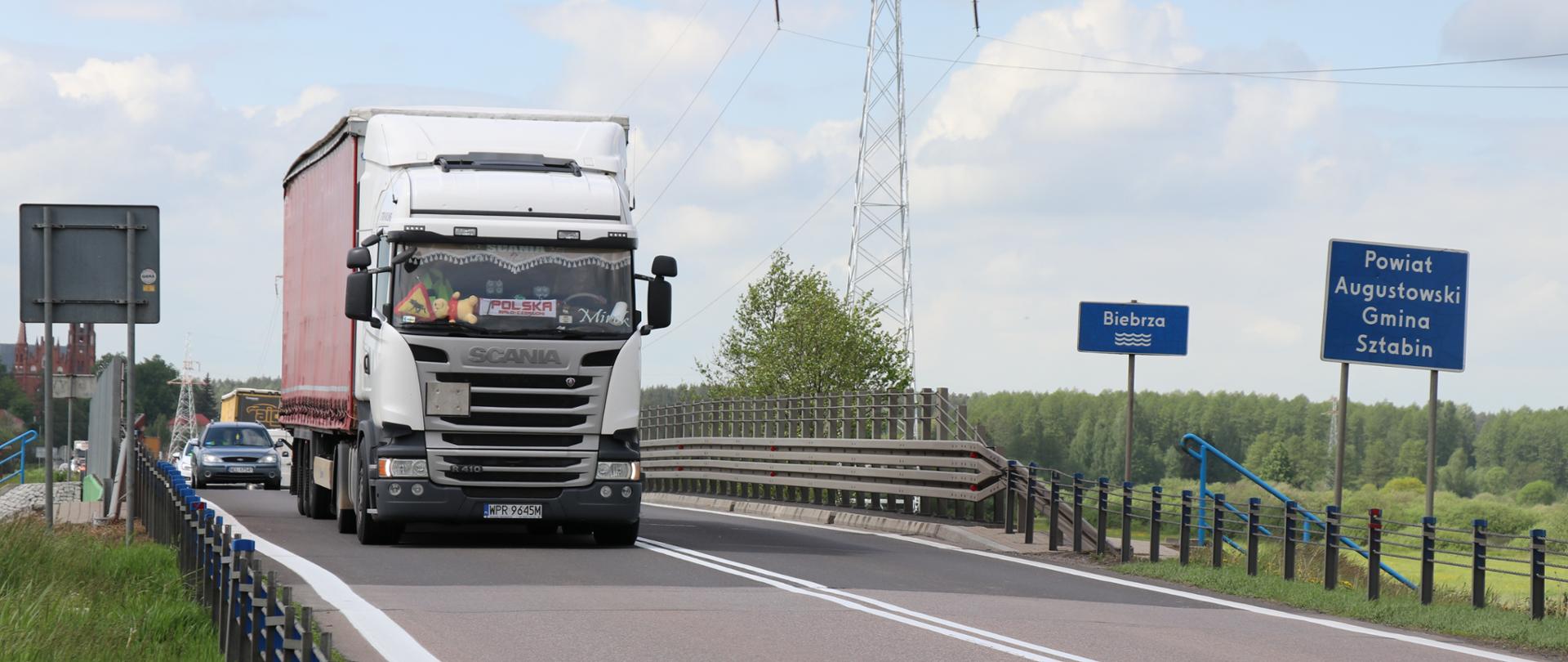 Po lewej stronie ciężarówka na moście nad rz. Biebrza, po prawej tablice z oznaczeniem rz. Biebrzy i granicy powiatu augustowskiego, gminy Sztabin. 