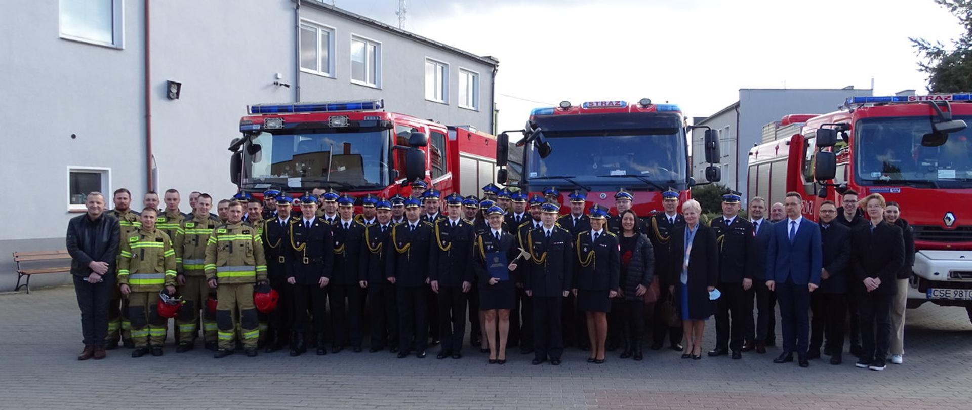 Zdjęcie wykonano w ciągu dnia na placu Komendy Powiatowej Państwowej Straży Pożarnej w Sępólnie Krajeńskim. Strażacy Państwowej Straży Pożarnej oraz goście pozują do zdjęcia na tle samochodów ratowniczo- gaśniczych. Na dalszym tle budynek Komendy Powiatowej Państwowej Straży Pożarnej. 