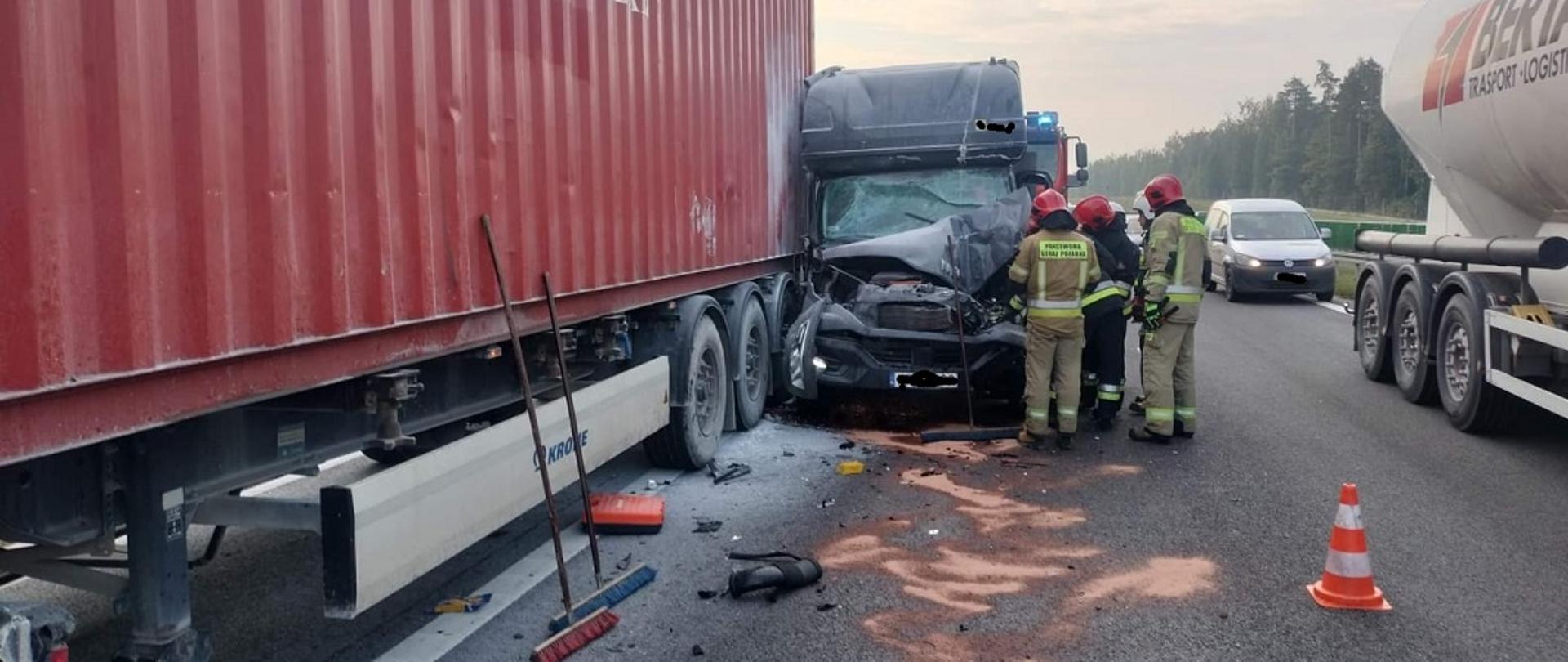 Na zdjęciu widoczna jest część naczepy samochodu ciężarowego, w którą uderzył dostawczy bus. Bus ma znacznie zdeformowaną kabinę kierowcy. Przy uszkodzonym busie stoi pięciu raowników straży pożarnej dwóch z nich ubranych jest w ubrania bojowe koloru żółtego pozostali koloru czarnego. Wszyscy ratownicy na głowach mają hełmy. Na jezdni widać rozlane substancje eksploatacyjne zabezpieczone sorbentem w postaci pomarańczowych plam. Na jezdni widoczne są również rozrzucone elementy z uszkodzonych pojazdów. Obok widoczna jest część naczepy cysterny oraz biały samochód typu pikap.