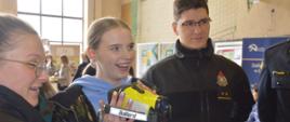 Targi edukacyjne. Sala sportowa w ZSZ w Rawiczu. Strażacy prezentują uczennicom kamerę termowizyjną. Jest ona trzymana przez jedną z dziewczyn. Ma kolor żółty.