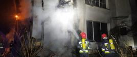 Na zdjęciu widzimy spaloną przybudówkę budynku mieszkalnego. Strażacy zabezpieczeni w aparaty dróg oddechowych podają prąd wody na żarzące się elementy konstrukcyjne przybudówki.