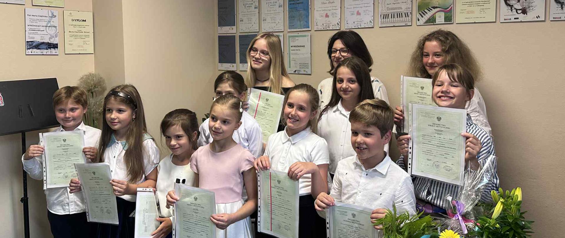 To zdjęcie przedstawia dużą grupę uczniów wraz z nauczycielką stojących w sali w szkole muzycznej. Jest to scena z zakończenia roku szkolnego, gdzie uczniowie odbierają dyplomy i nagrody. Na ścianie za nimi wisi wiele oprawionych dyplomów i certyfikatów, co potwierdza, że jest to miejsce związane z edukacją i osiągnięciami. Na pierwszym planie, od lewej do prawej, widać: Młodego chłopca w białej koszuli i ciemnych spodniach, uśmiechniętego i trzymającego w rękach dyplom. Dziewczynkę w białej bluzce z długim rękawem i ciemnej spódnicy, trzymającą dyplom. Dziewczynkę w białej bluzce z krótkim rękawem i niebieskiej spódnicy, również z dyplomem. Dziewczynkę w jasnej sukience, uśmiechniętą, trzymającą dyplom. Kolejną dziewczynkę w białej bluzce i ciemnej spódnicy, uśmiechniętą, trzymającą dyplom. Z przodu po prawej stronie, chłopca w białej koszuli, uśmiechniętego i trzymającego dyplom. Tuż za nim, dziewczynkę w jasnej bluzce i ciemnej spódnicy, trzymającą dyplom. Dalej w prawym rogu, widać również bukiety kwiatów i ozdobne paczki, prawdopodobnie prezenty dla uczniów lub nauczycieli. Za nimi, w drugim rzędzie, stoi więcej uczniów i dorosłych: Kilka młodych dziewcząt, w tym jedna w okularach, trzymające dyplomy. Dorosła kobieta w okularach, uśmiechnięta, patrząca w stronę dzieci.
Wszyscy na zdjęciu wydają się być zadowoleni i dumni ze swoich osiągnięć, co oddaje radosną atmosferę zakończenia roku szkolnego.