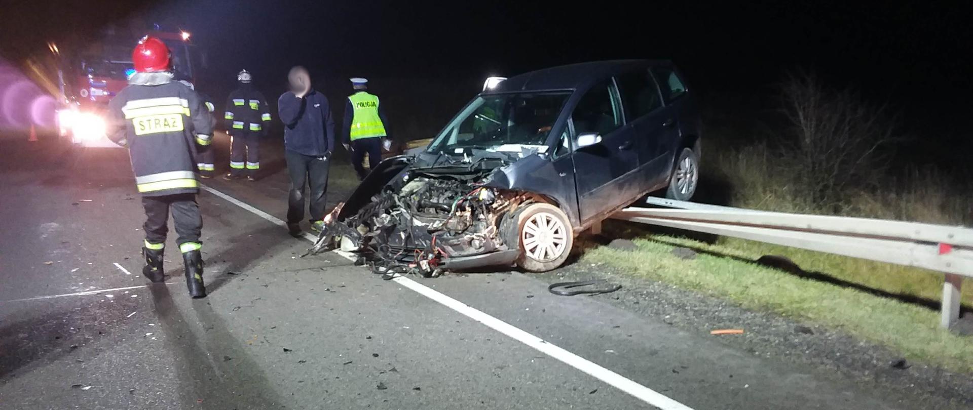 Pora nocna, wrak samochodu osobowego po zderzeniu, zarzucony tyłem na barierkę energochłonną . Widoczne rozbite elementy samochodu i karoserii, przy samochodzie Strażacy i Policjant prowadzący swoje czynności. 