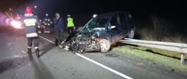 Pora nocna, wrak samochodu osobowego po zderzeniu, zarzucony tyłem na barierkę energochłonną . Widoczne rozbite elementy samochodu i karoserii, przy samochodzie Strażacy i Policjant prowadzący swoje czynności. 